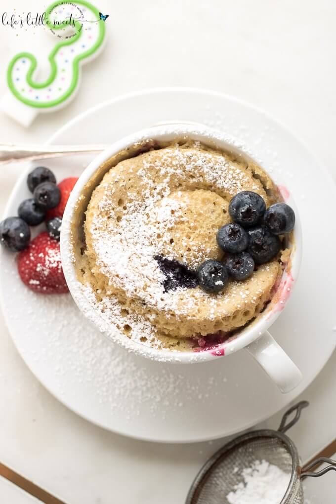 overhead view of this mug cake with berries on top and on the plate with a white background