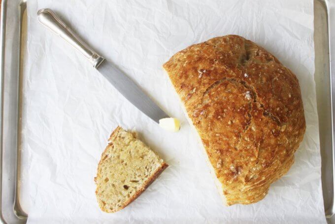 No-Knead Oatmeal Bread cut in half with a buttered slice and a bread knife on a rimmed baking sheet with parchment paper