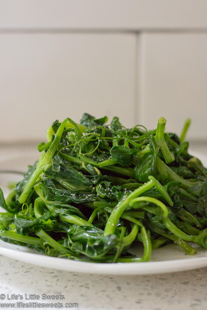 Sautéed Pea Shoots piled on a white plate with white subway tile in the background