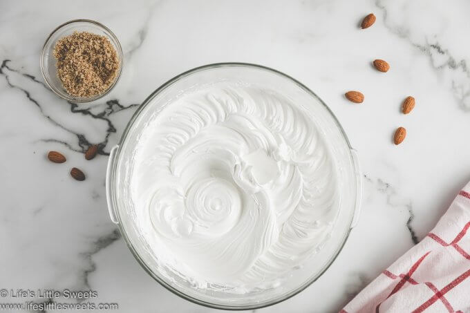 whipped egg whites in a clear glass bowl with added almond extract