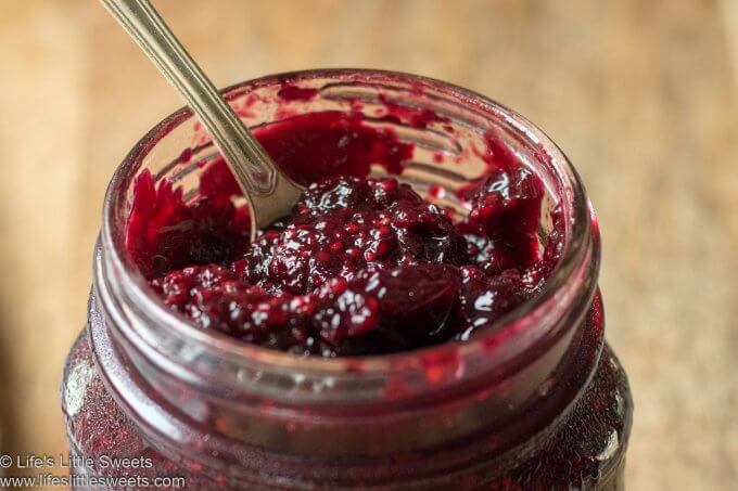 a clear glass jar of jam with a spoon in it with a wood background