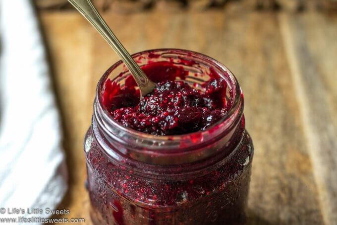Red jam in a mason jar with a spoon on a wood cutting board with a white tea towel
