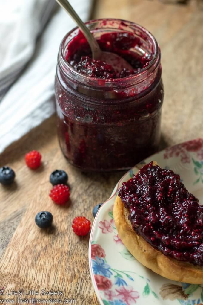 Red jam in a mason jar with a spoon on a wood cutting board with a white tea towel and jam toast on a colorful matching plate