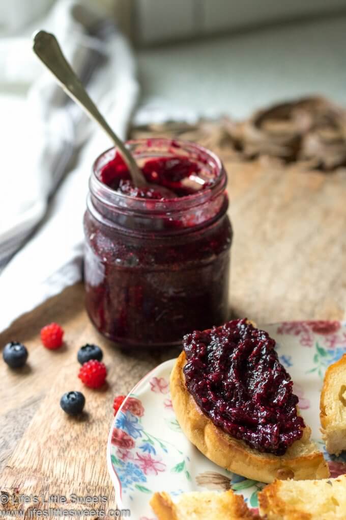 Red jam in a mason jar with a spoon on a wood cutting board with a white tea towel with toasted bagels and jam and fresh berries