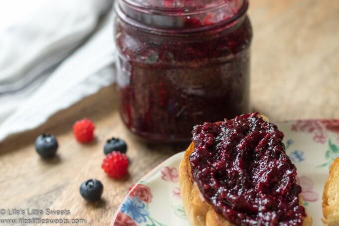 red jam in a mason jar with fresh berries and more jam on a buttered, toasted bagel all on a wood cutting board with a white tea towel in the background