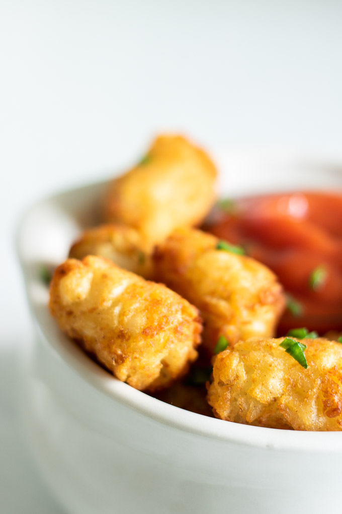 a close up of tater tots in a white bowl with green chives on top and ketchup in the bowl