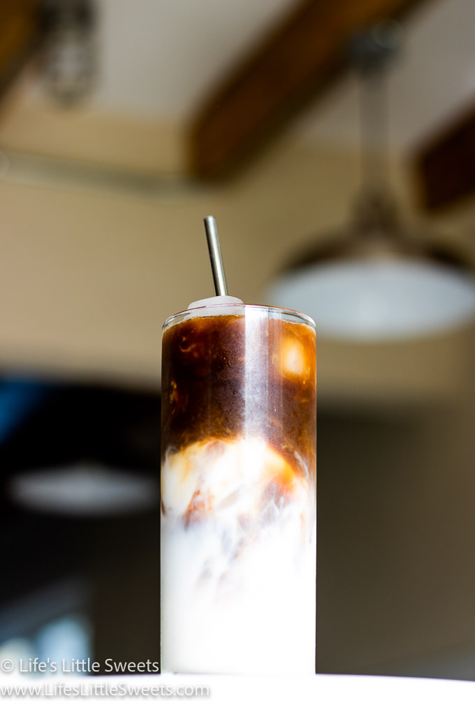 Iced Coffee with Almond Milk on a marble counter with a stainless steel straw with stainless steel pendant ceiling lamps in the background