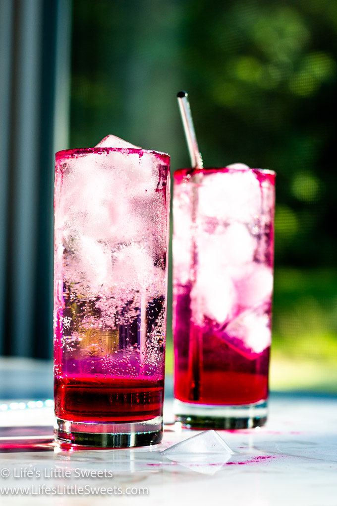 2 pink soda drinks, 1 with a metal straw, on a white table near a window