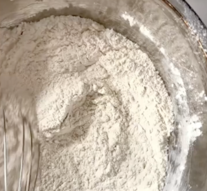 a metal mixing bowl, filled with a flour mixture being whisked with a metal whisk