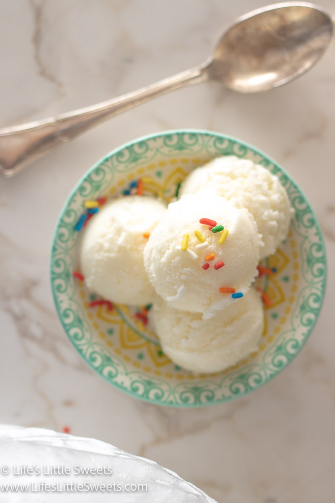 scoops of plain vanilla ice cream with sprinkles in a shallow colorful dish on a white marble surface with a silver spoon