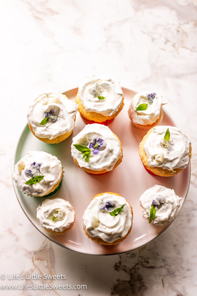 a white cake stand with 9 angel food cupcakes, small and regular-sized, topped with a white frosting, small, green mint leaves and sugared white and purple violets