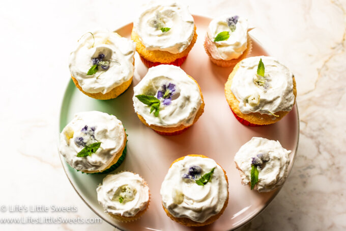 9 light-colored cupcakes decorated with mint leaves and sugared flowers, large and small on a white cake stand over a white marble table surface