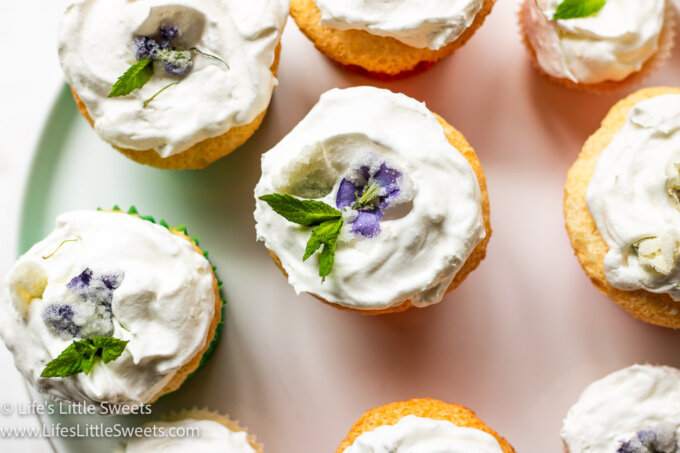 a white plate that has several light-colored cupcakes on it, decorated with mint leaves and sugared flowers