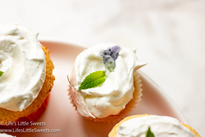 light-colored cupcakes with a white frosting topping and decorated with mint leaves and sugared violets on a white plate over a marble surface