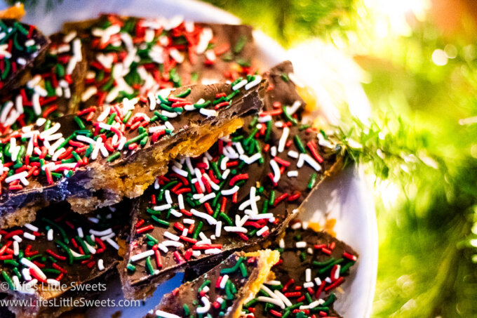 horizontal photo of a white plate filled with Christmas Chocolate Toffee near a white-lighted Christmas tree