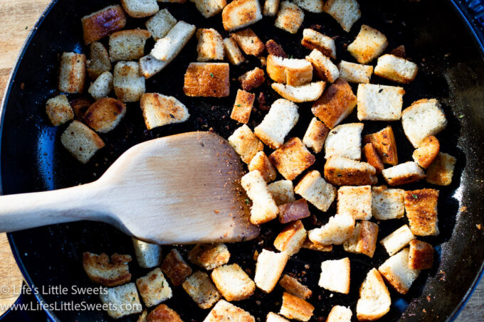 croutons being toasted in a black pan with a wooden spatula